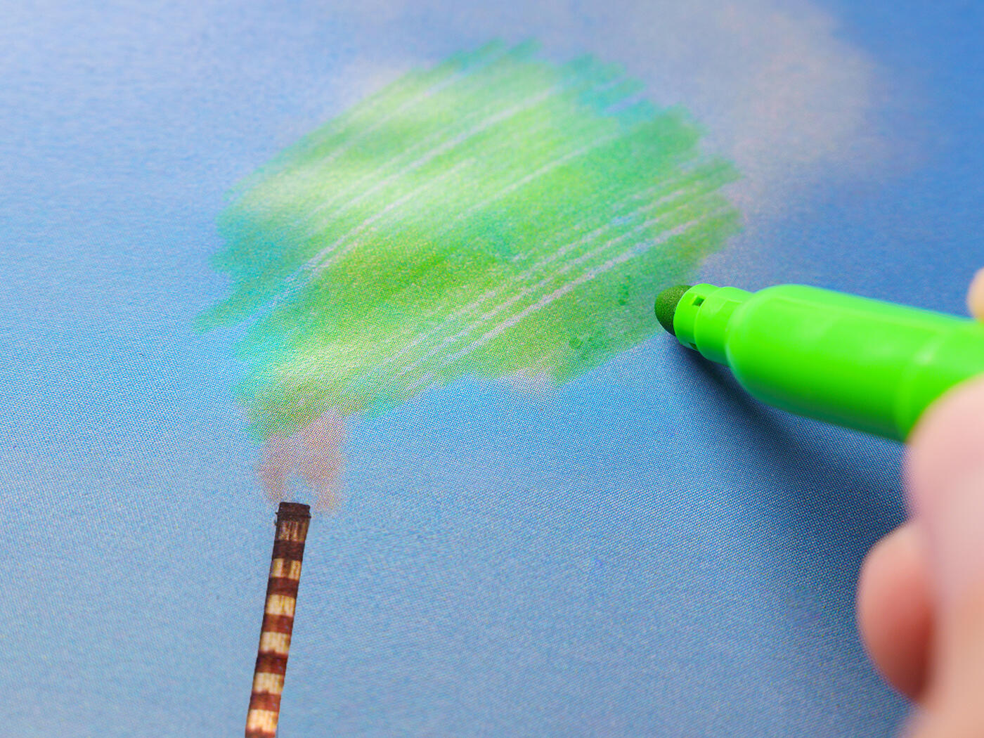 A hand colors over smoke from a factory chimney with green marker