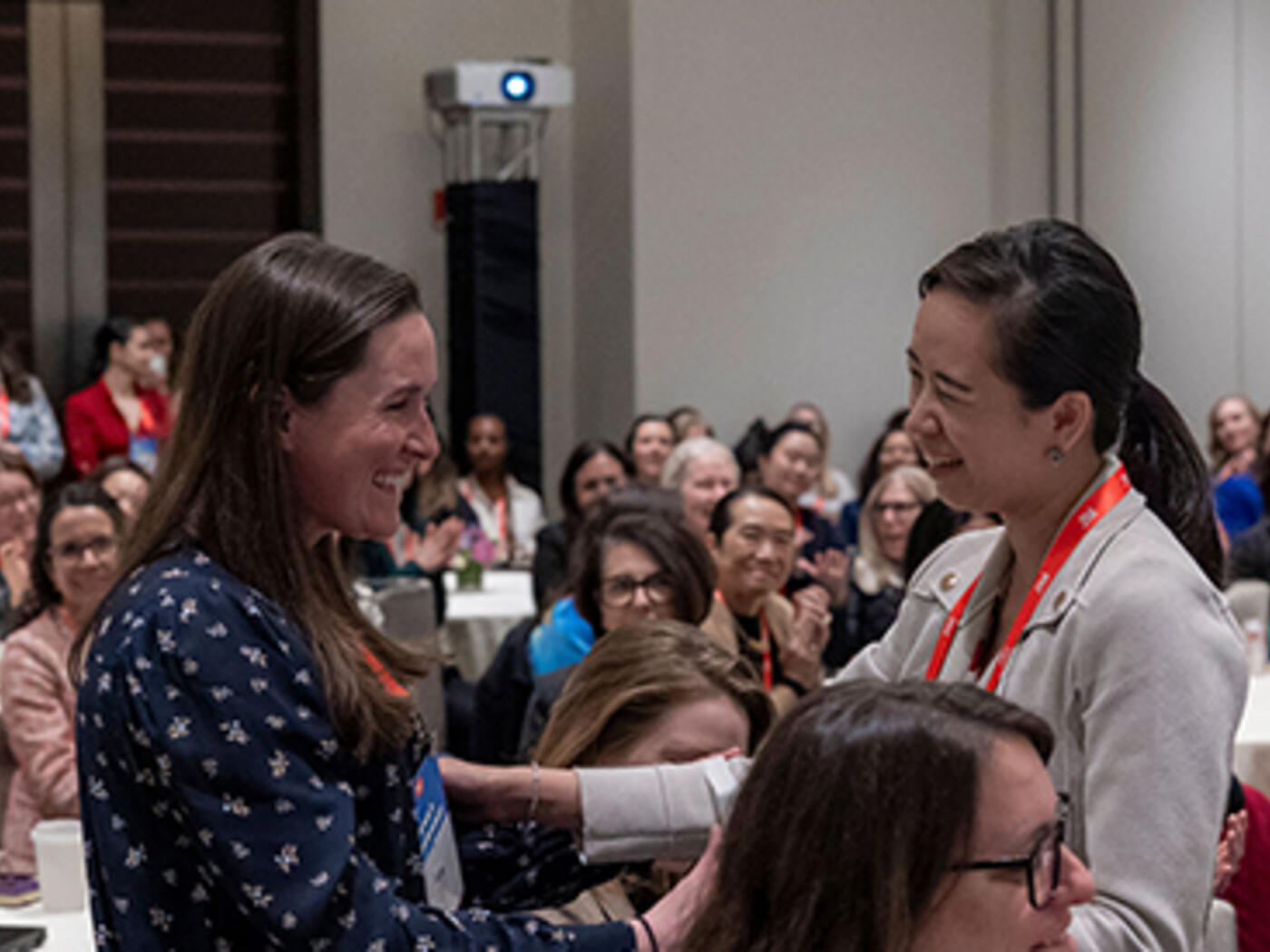 Two women embrace in the foreground of a packed room at a conference
