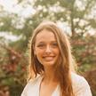 A photo of Nora Donnelly, wearing a white top, smiling, outside, in front of a blurred background of trees